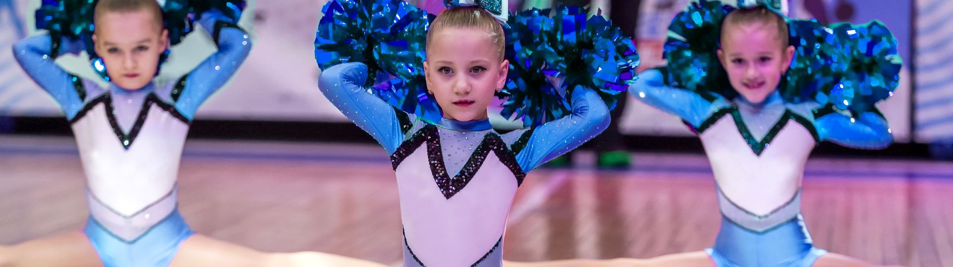Three young cheerleaders in blue and white outfits are smiling, holding blue pom-poms above their heads while sitting on the floor.