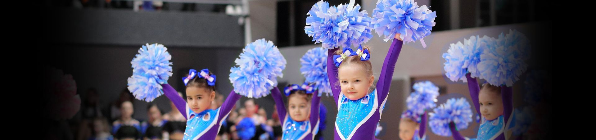 Young cheerleaders in matching purple and blue outfits raise pom-poms. They perform indoors with smiling faces.