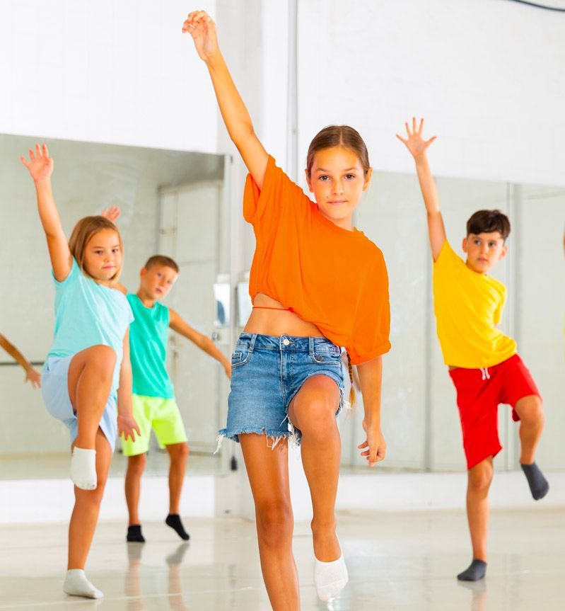 Children dancing in a studio. A girl in orange raises an arm, others in colorful shirts and shorts also dance.