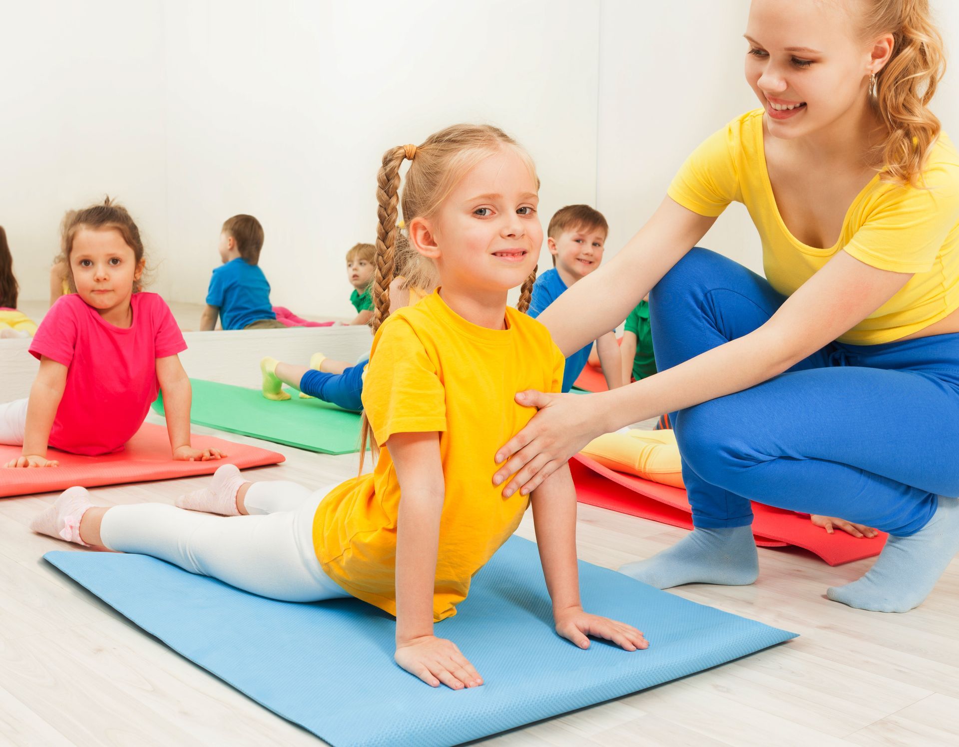 Children in a yoga class doing the cobra pose; instructor assists a girl in a yellow shirt.
