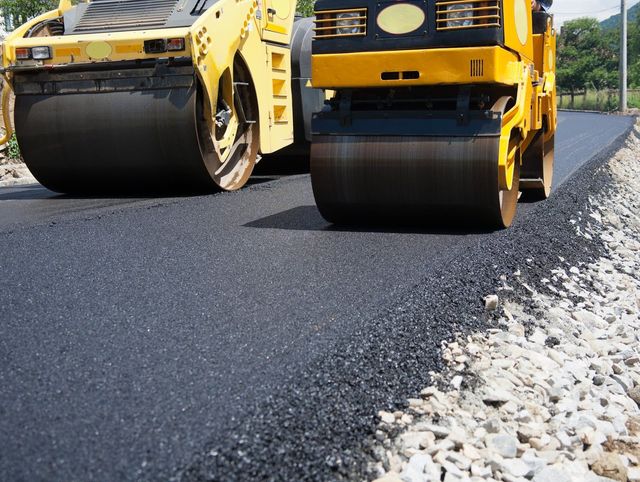 Two yellow road rollers paving fresh asphalt beside a gravel shoulder