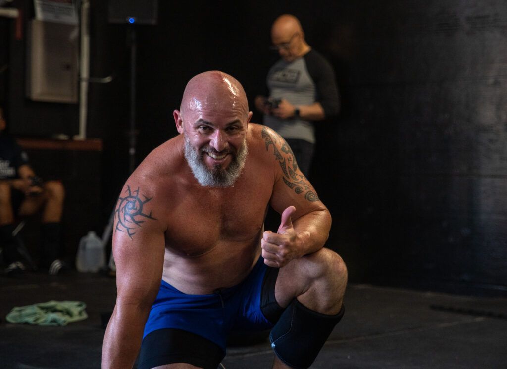 A shirtless man with a beard is squatting down in a gym and giving a thumbs up.