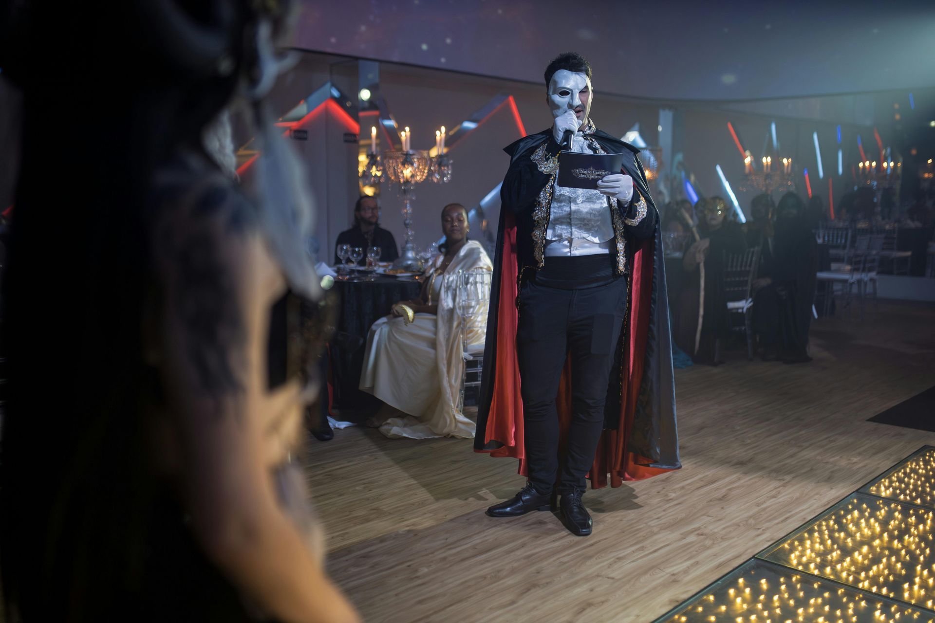 Man in costume speaks at event, red cape, lights.