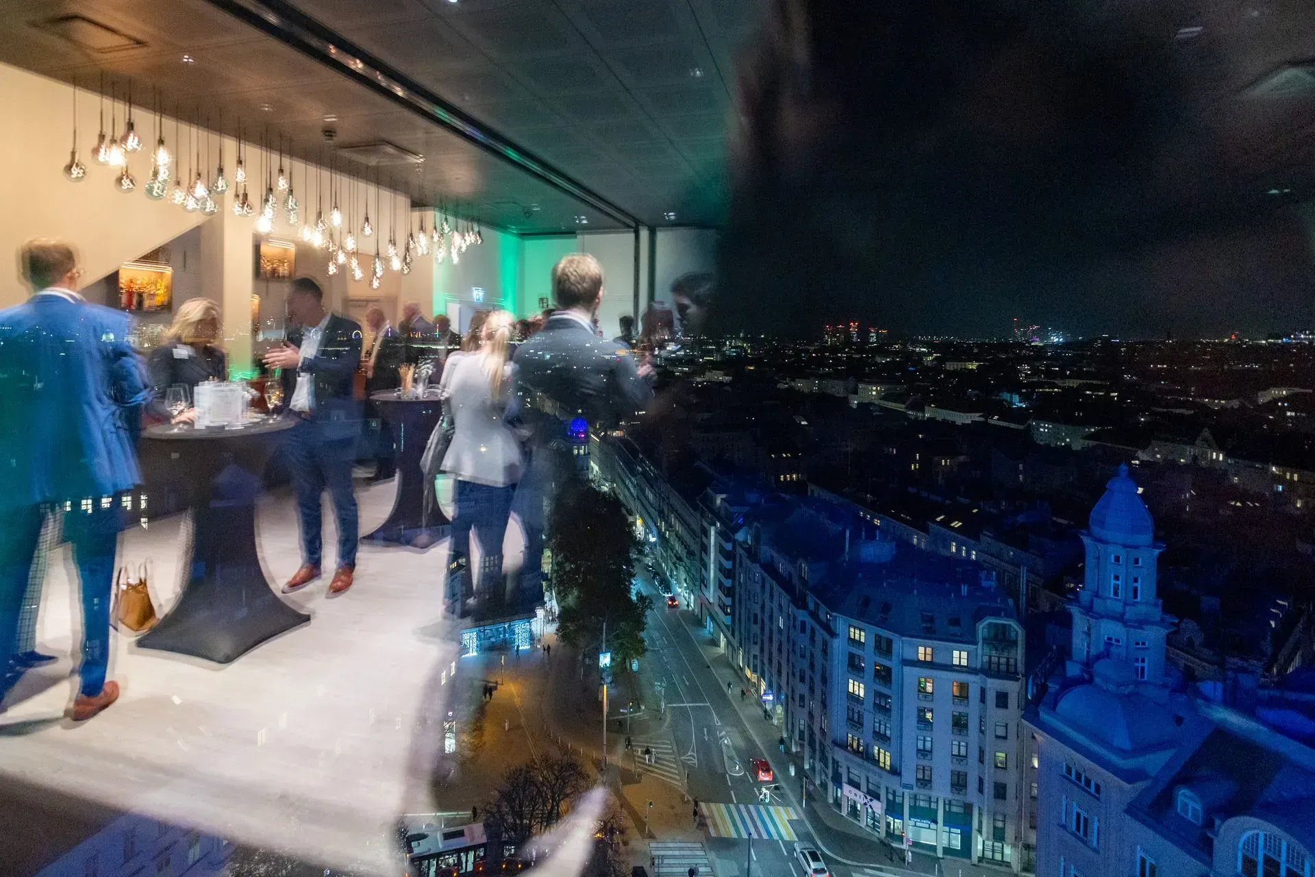 Eine Gruppe von Menschen steht in einem Raum um Tische herum und bietet Ausblick auf eine Stadt bei Nacht.