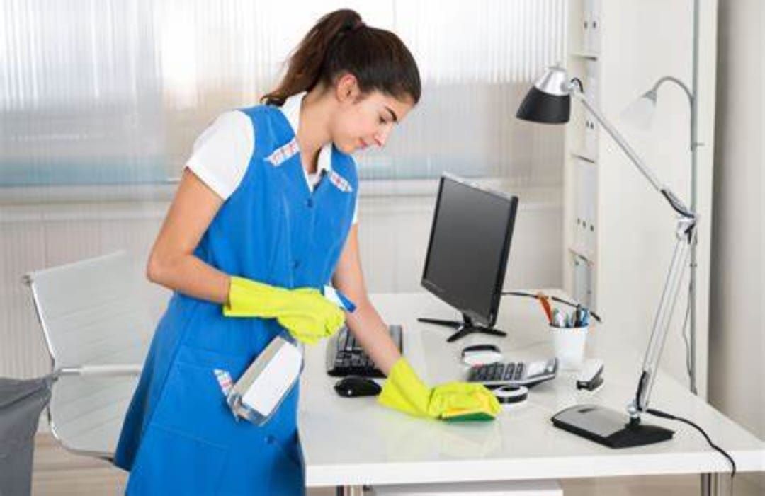 Person wearing blue uniform and yellow gloves cleaning an office desk.