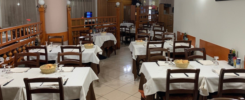 Sala da pranzo vuota di un ristorante con tovaglie bianche, sedie di legno e tavoli apparecchiati con cura.