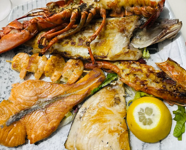 Piatto misto di frutti di mare grigliati con salmone, aragosta, pesce e guarnizione di limone su un piatto bianco.