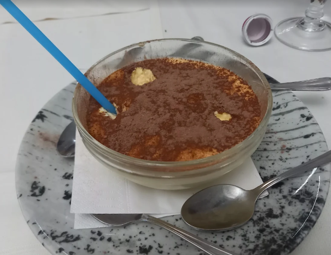 Una ciotola di vetro con tiramisù e cacao in polvere, adagiata su un piatto, con un cucchiaio accanto.
