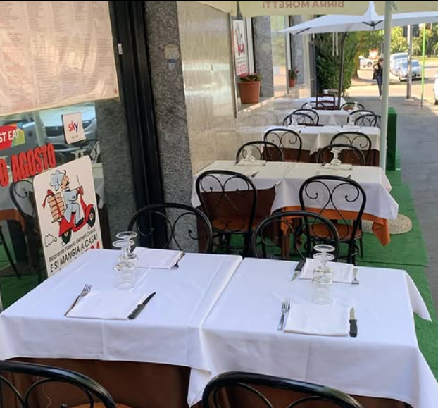 Tavolini da caffè all'aperto con tovaglie bianche e sedie nere lungo un marciapiede di fronte a un negozio.