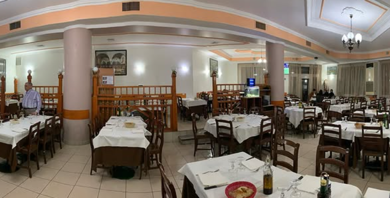 Sala da pranzo vuota di un ristorante con tovaglie bianche, sedie di legno e luci calde sul soffitto.