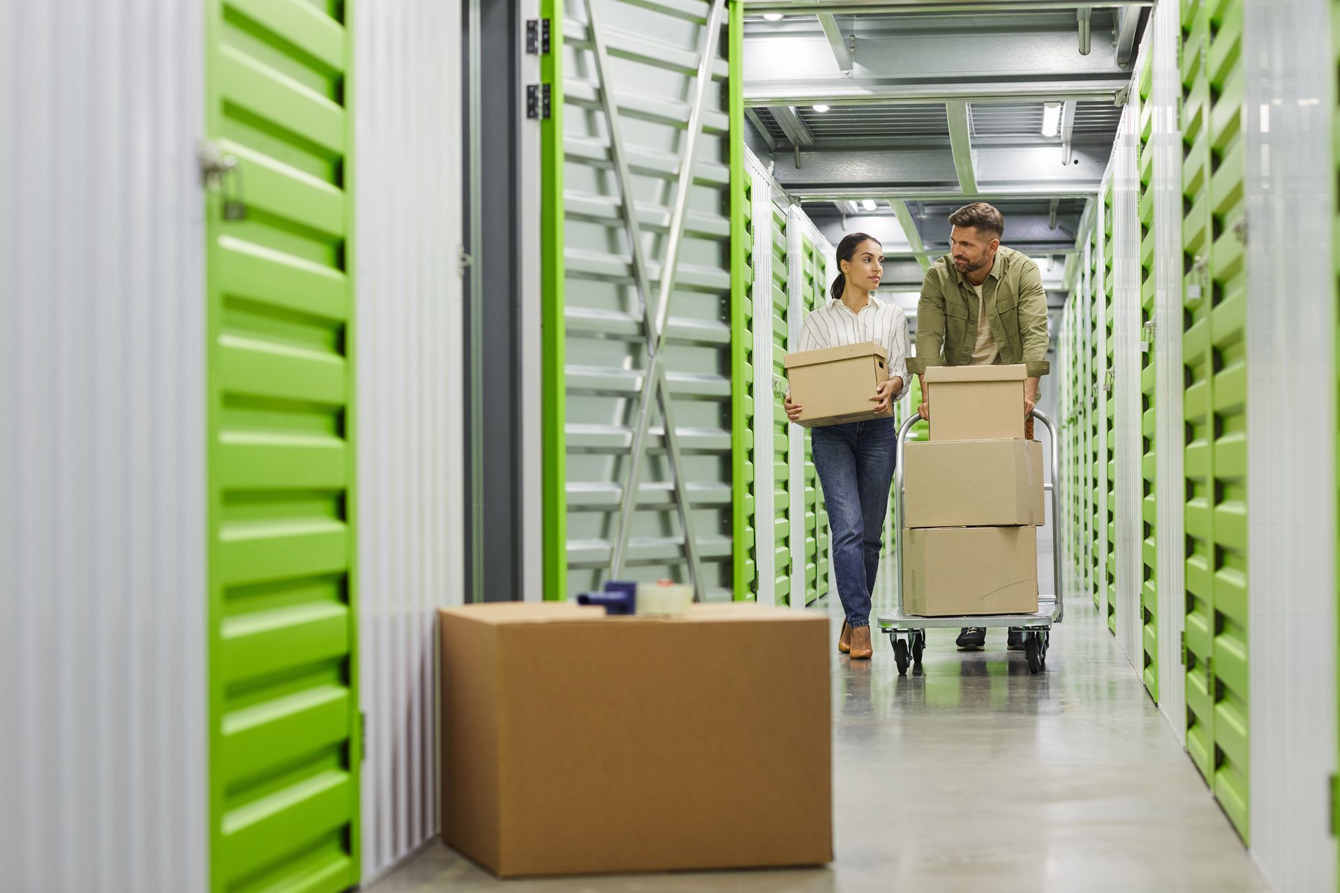 A young couple holding cardboard boxes walking in a hallway with green doors to a storage unit.