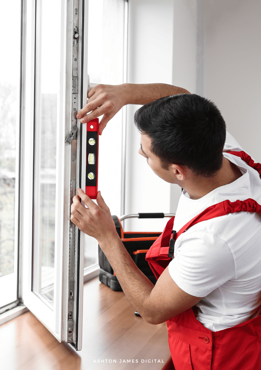 Man using a level to install a window in a white-walled room, tool bag nearby.