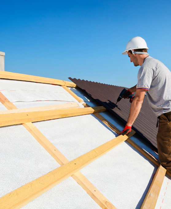 Roofer in white hard hat uses a nail gun on a roof with wooden beams and white insulation under a blue sky.