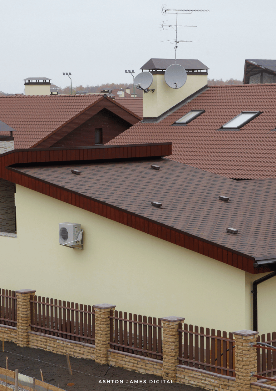 Roofs of a residential building with brown shingles, chimney, satellite dishes, and a fence.