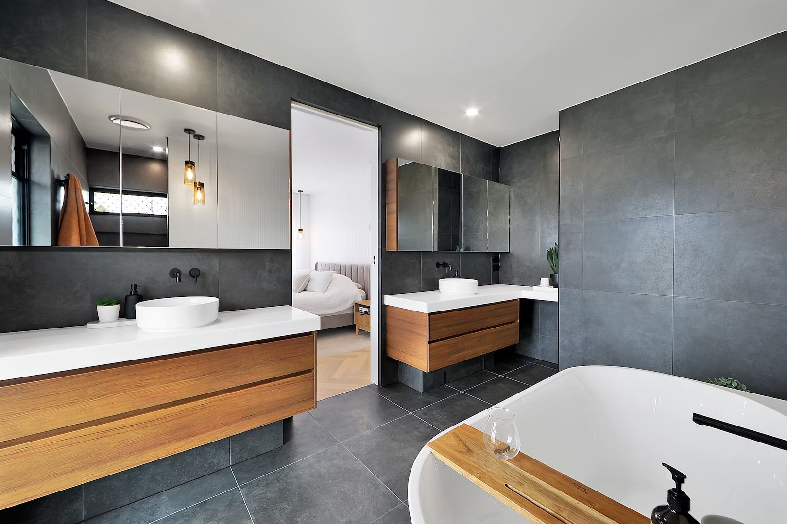 Modern bathroom with wooden vanities, white sinks, and a freestanding tub. Dark gray walls and floor.