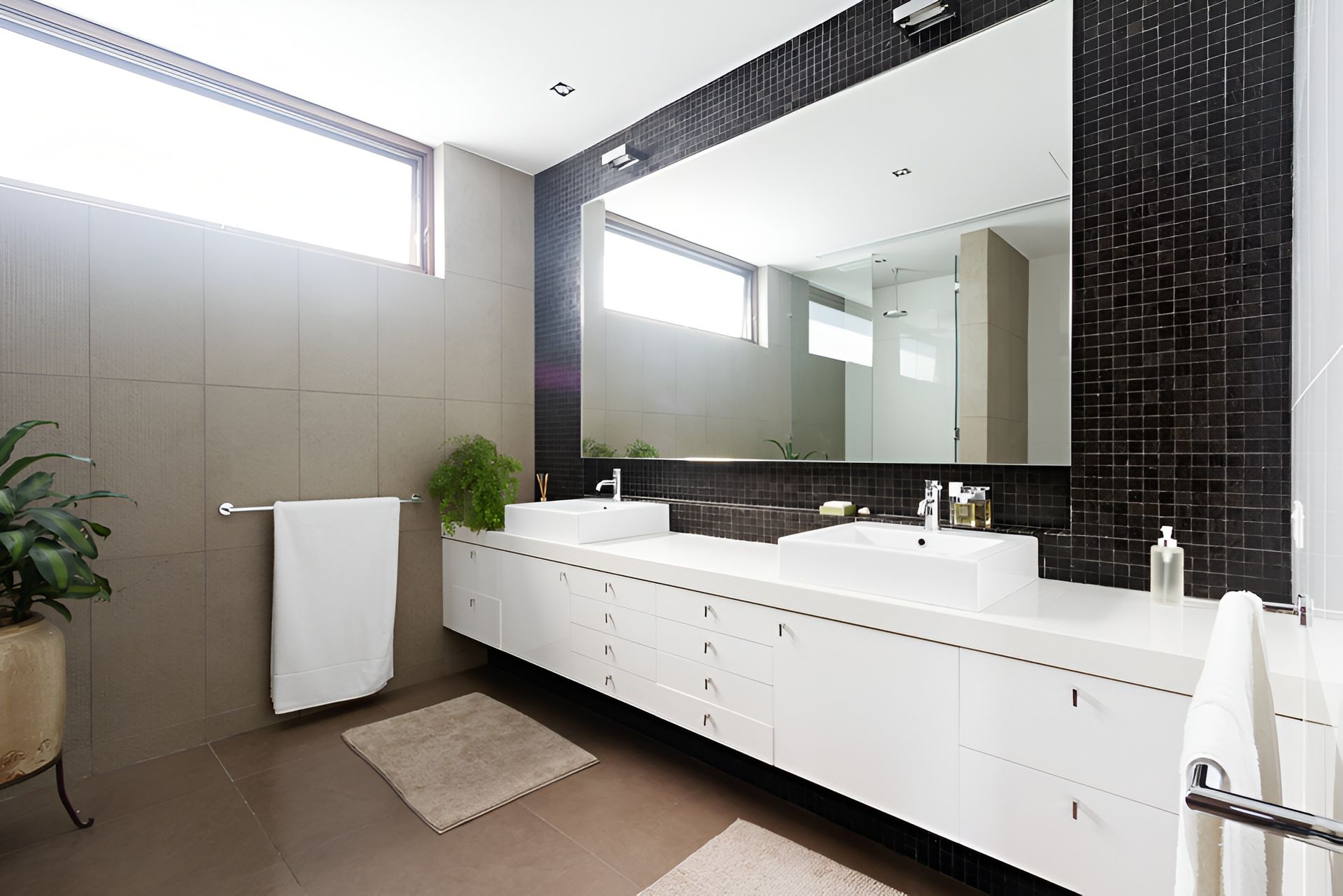 Modern bathroom with large mirror, double sinks, and white cabinets. Brown tile floor, black mosaic accent wall, and long window.