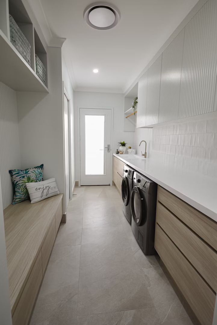 Laundry room with light wood cabinets, washer/dryer, and a bench seat. White walls, decorative tile, and a door.