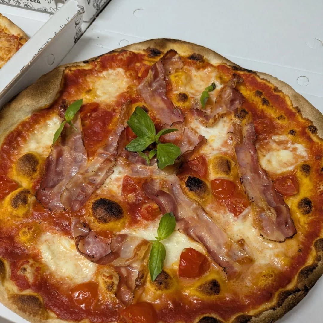 Pizza con pancetta, pomodori e basilico su un piatto bianco; scatola della pizza sullo sfondo.
