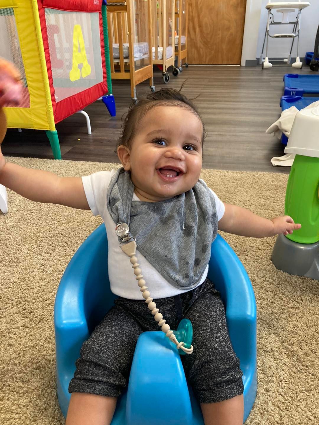 A baby is sitting in a blue chair with his arms outstretched and smiling.
