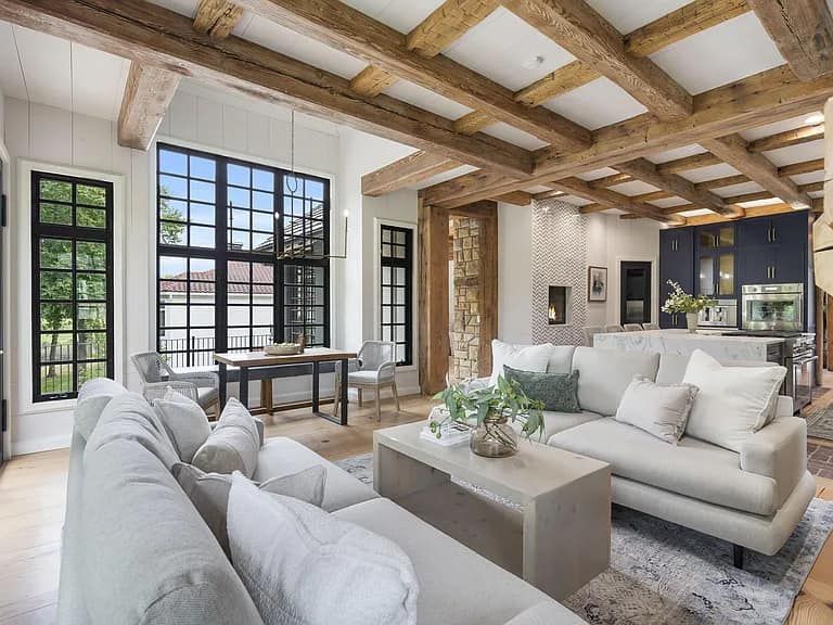 Living room with white sofas, wooden beams, large windows, and open kitchen.