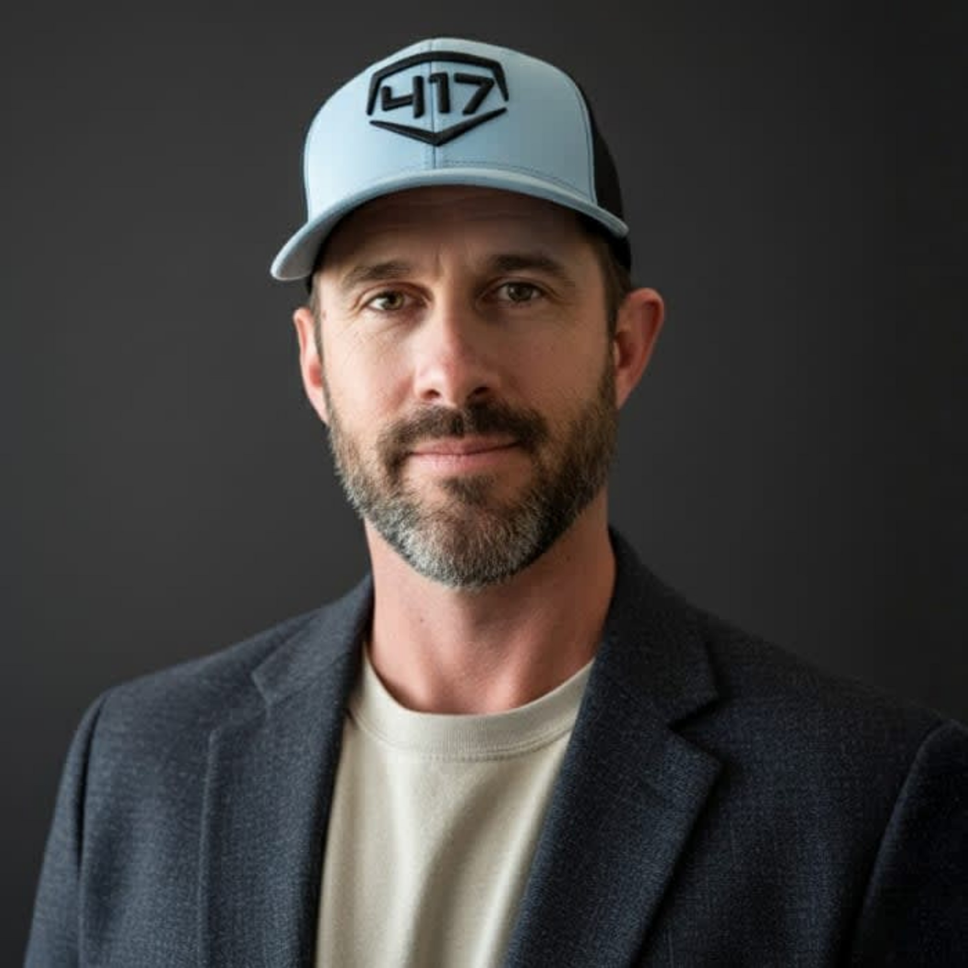 Man in baseball cap and blazer, looking at the camera.