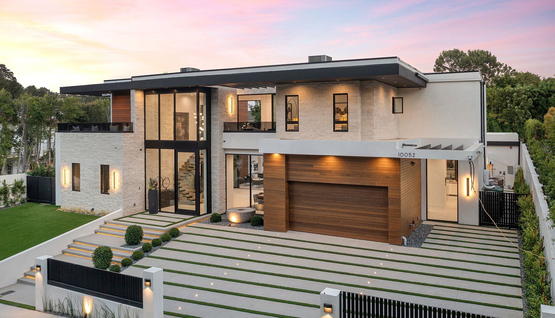 Modern two-story house with stone facade, glass walls, and wooden garage door, driveway with greenery, under a sunset sky.