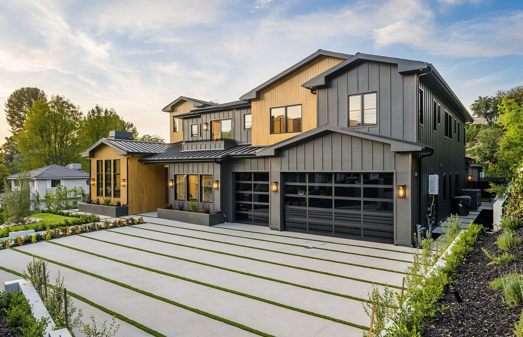 Modern two-story house with gray and wood siding, glass garage doors, and a long driveway lined with grass.