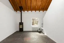 Stanza vuota con camino nero, finestra e soffitto in legno. Pareti bianche e pavimento in cemento.