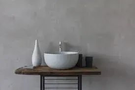 Lavandino bianco su una mensola in legno rustico contro una parete grigia testurizzata, con vaso decorativo e oggetti neri.