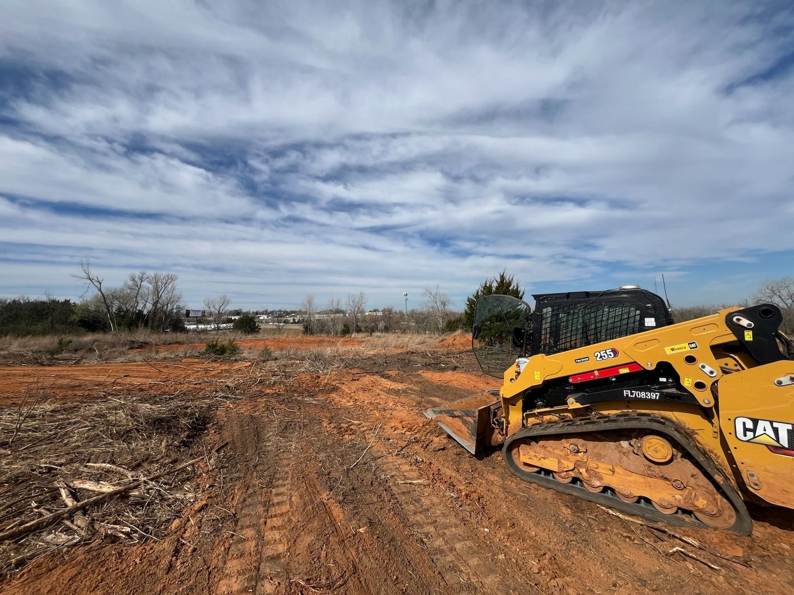 A yellow Caterpillar skid steer loader sits on a cleared, red-dirt construction site under a bright, cloudy sky.