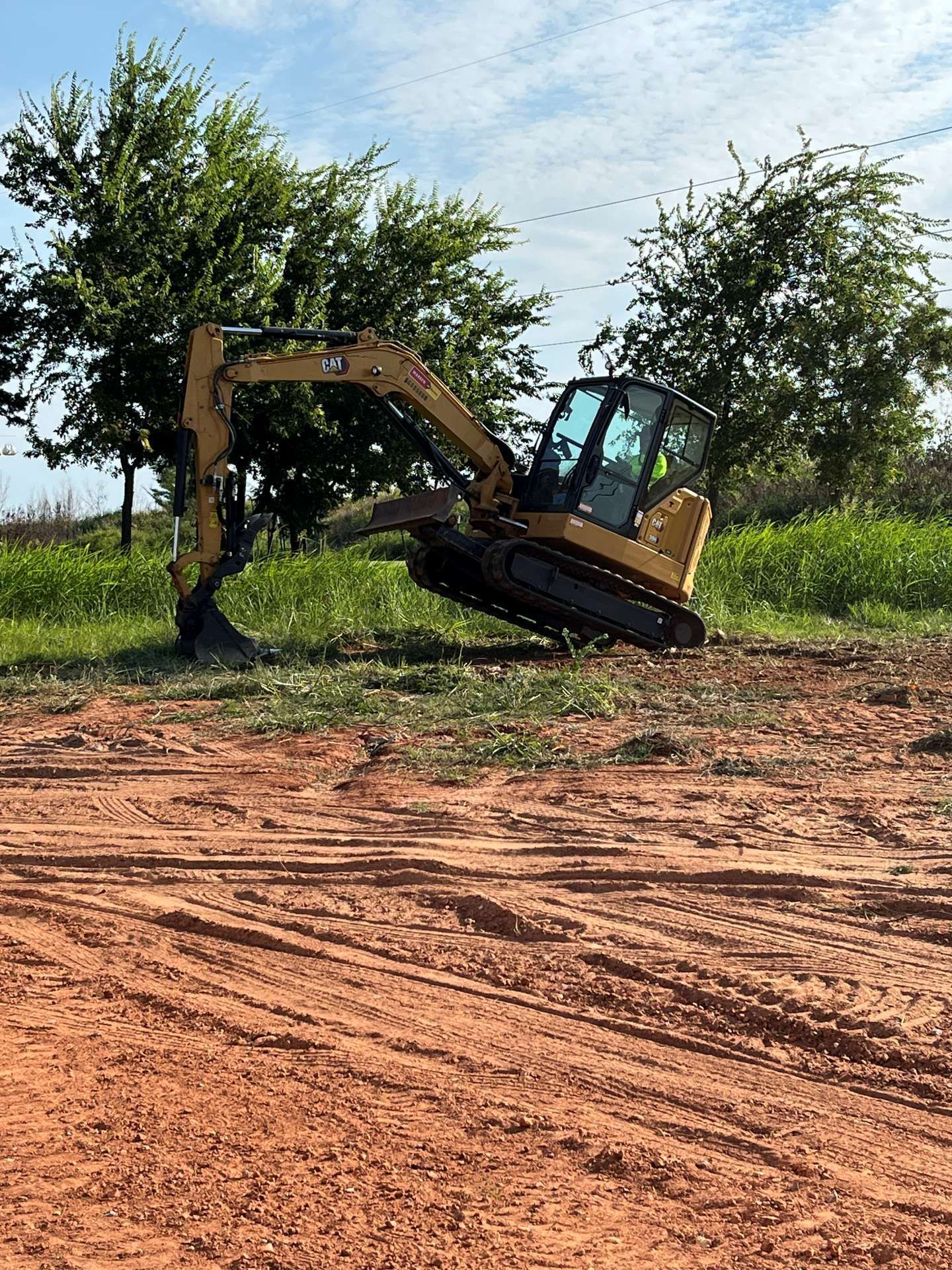 A yellow Caterpillar mini excavator sits on a dirt mound in a grassy field under a sunny, partly cloudy sky.