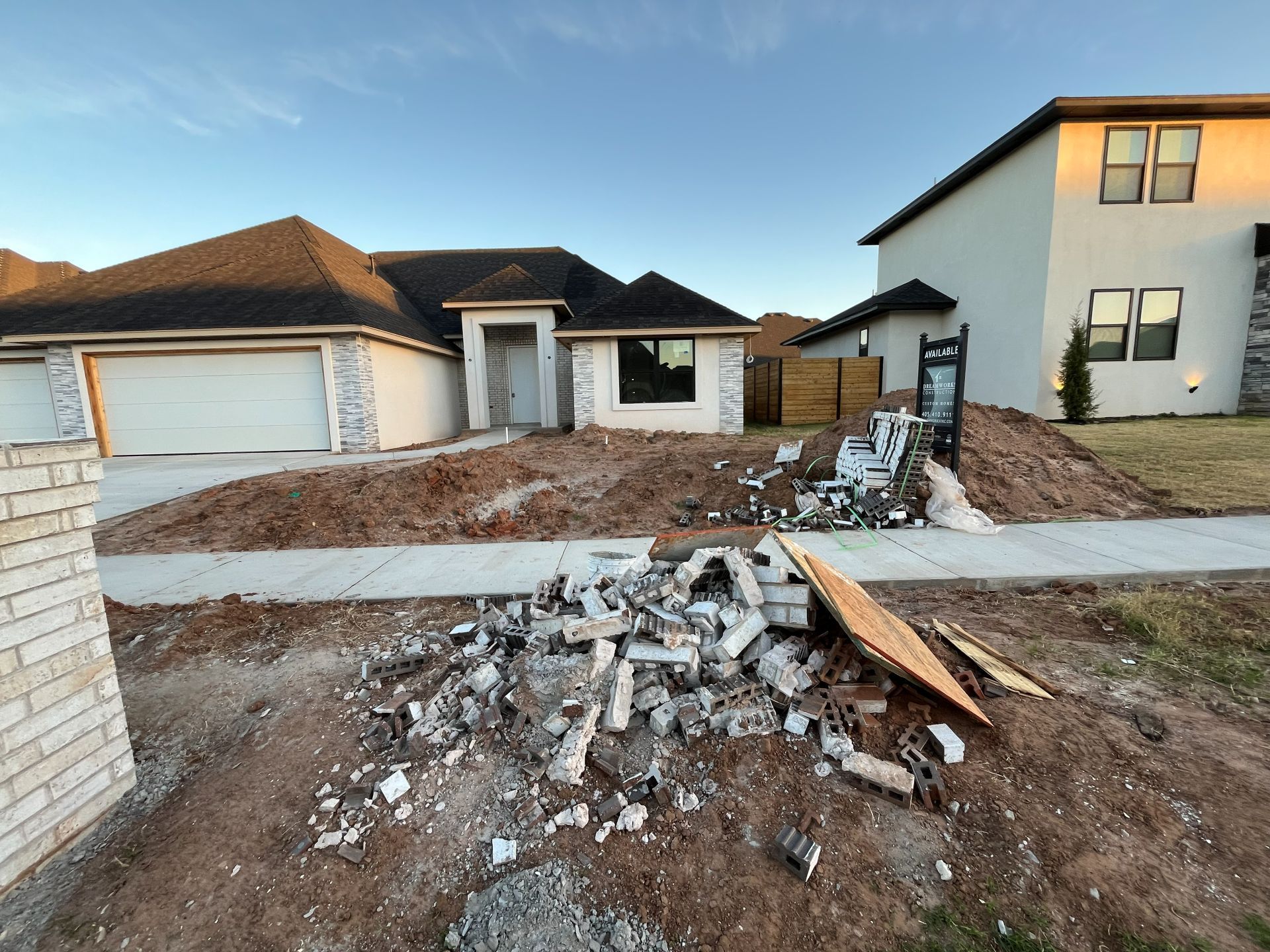A pile of construction debris sits on a dirt lot in front of a newly built house at sunset.