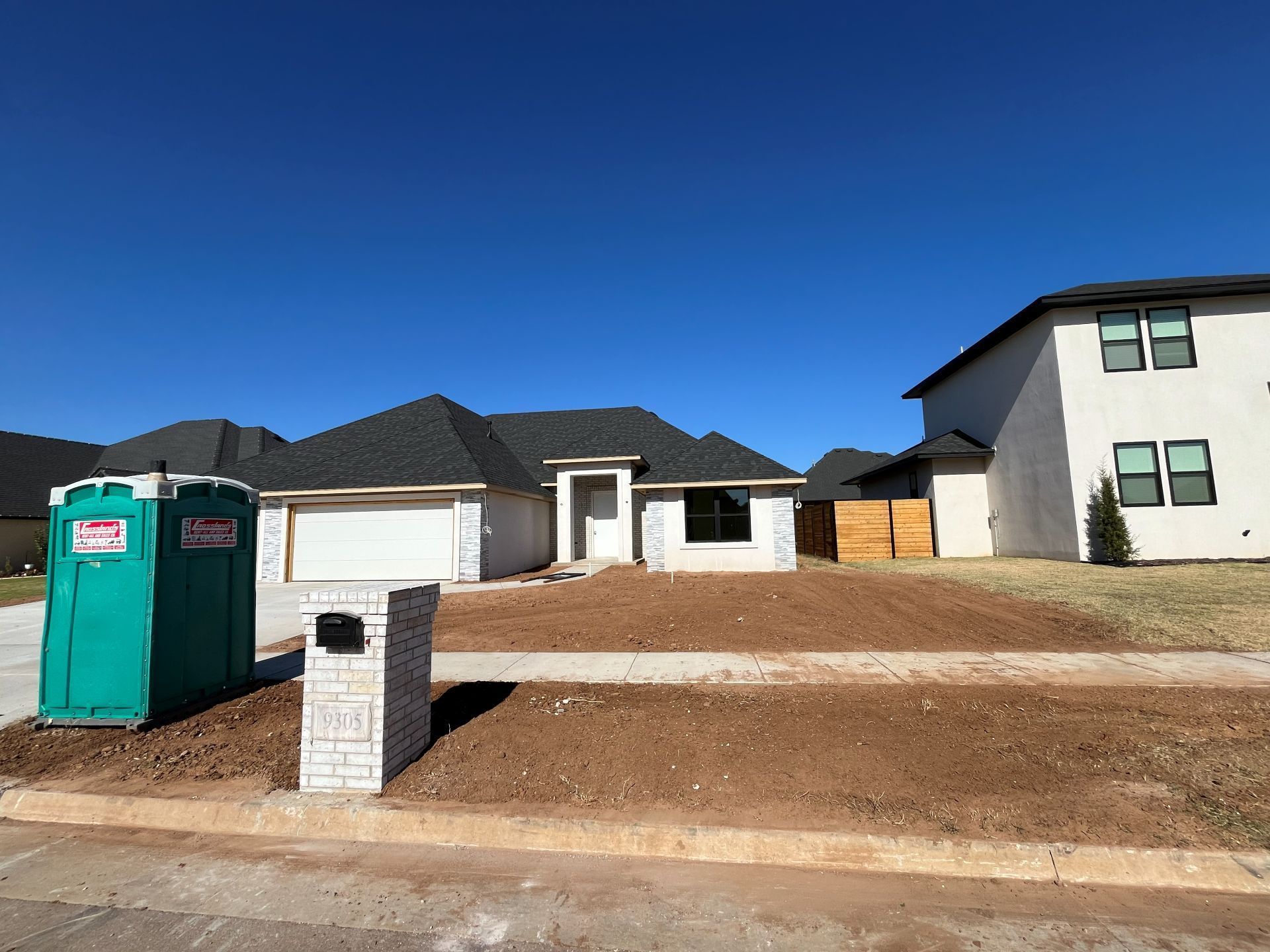 A new single-story house with a bare dirt front yard and a portable toilet on the lawn under a clear blue sky.
