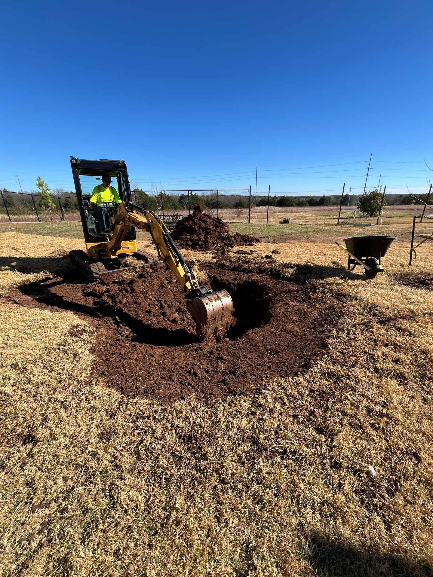 A person in a neon shirt operates a yellow excavator to dig a hole in a dry, grassy field under a clear blue sky.
