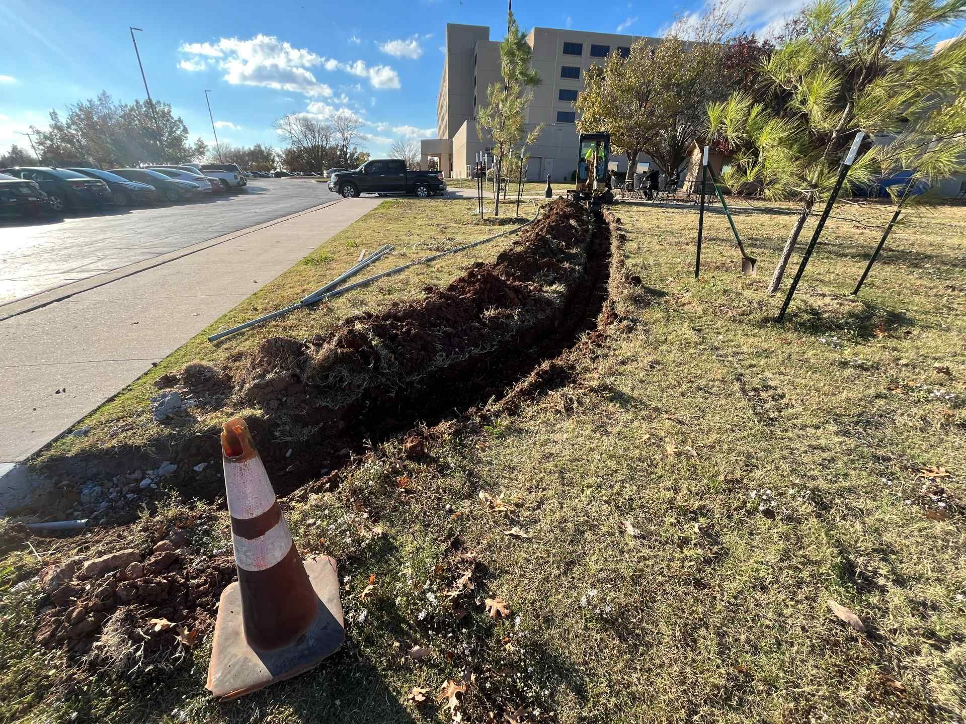 A trench dug into the grass next to a sidewalk and parking lot, marked by an orange traffic cone.