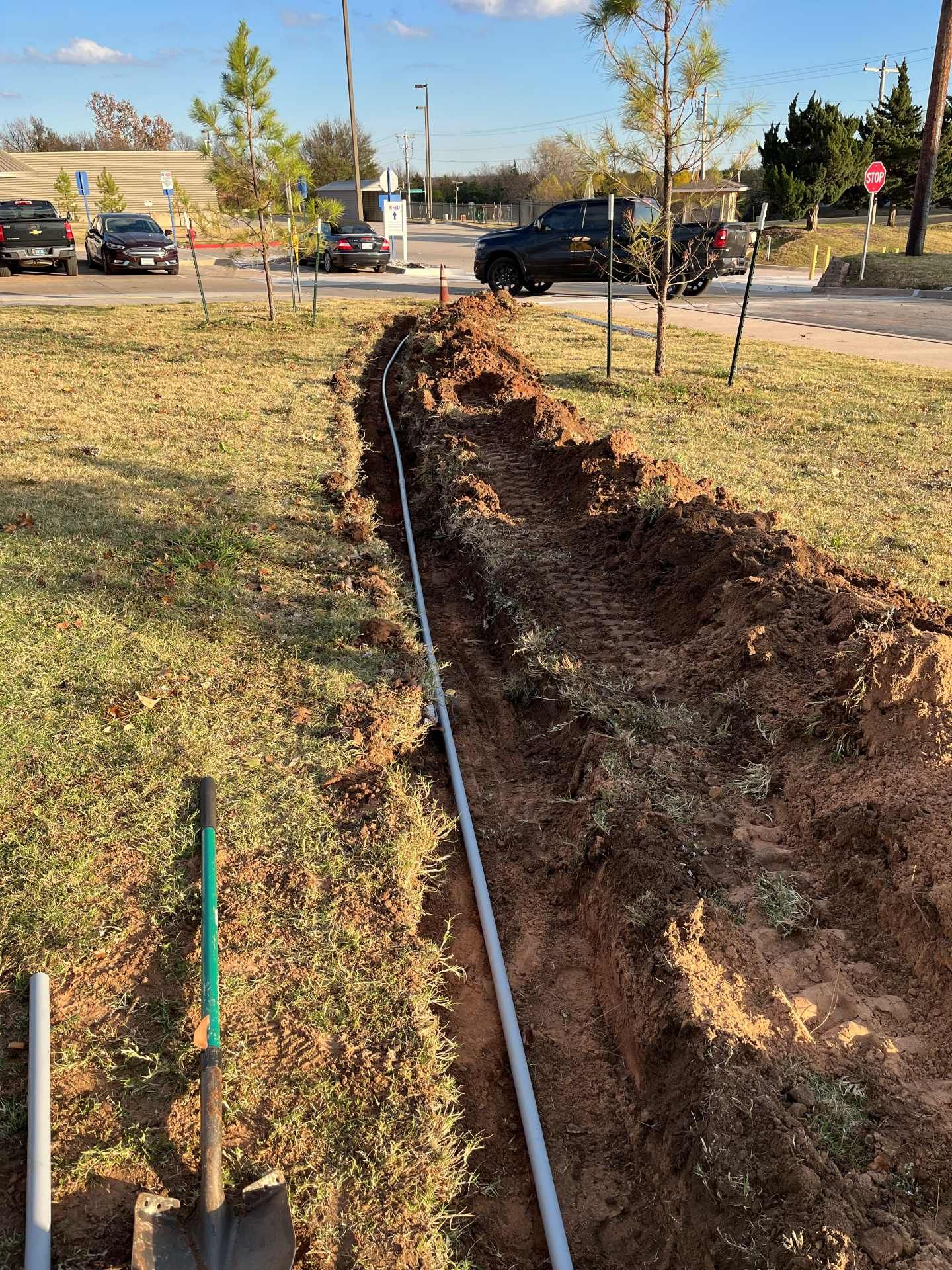 A long trench dug into a grass field with a gray irrigation pipe running through it and a shovel resting nearby.