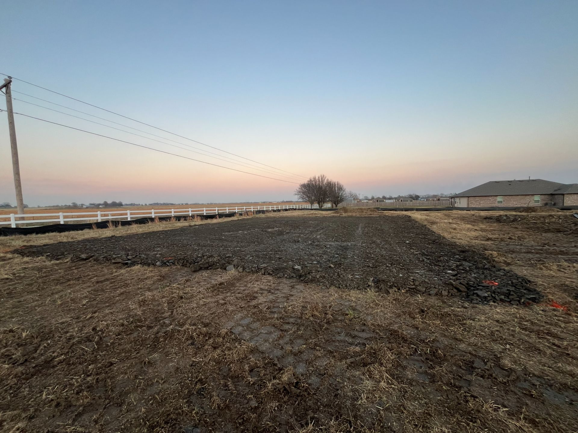 A rectangular patch of tilled, dark soil sits in a grassy field under a pale sunset sky with a distant house and tree.