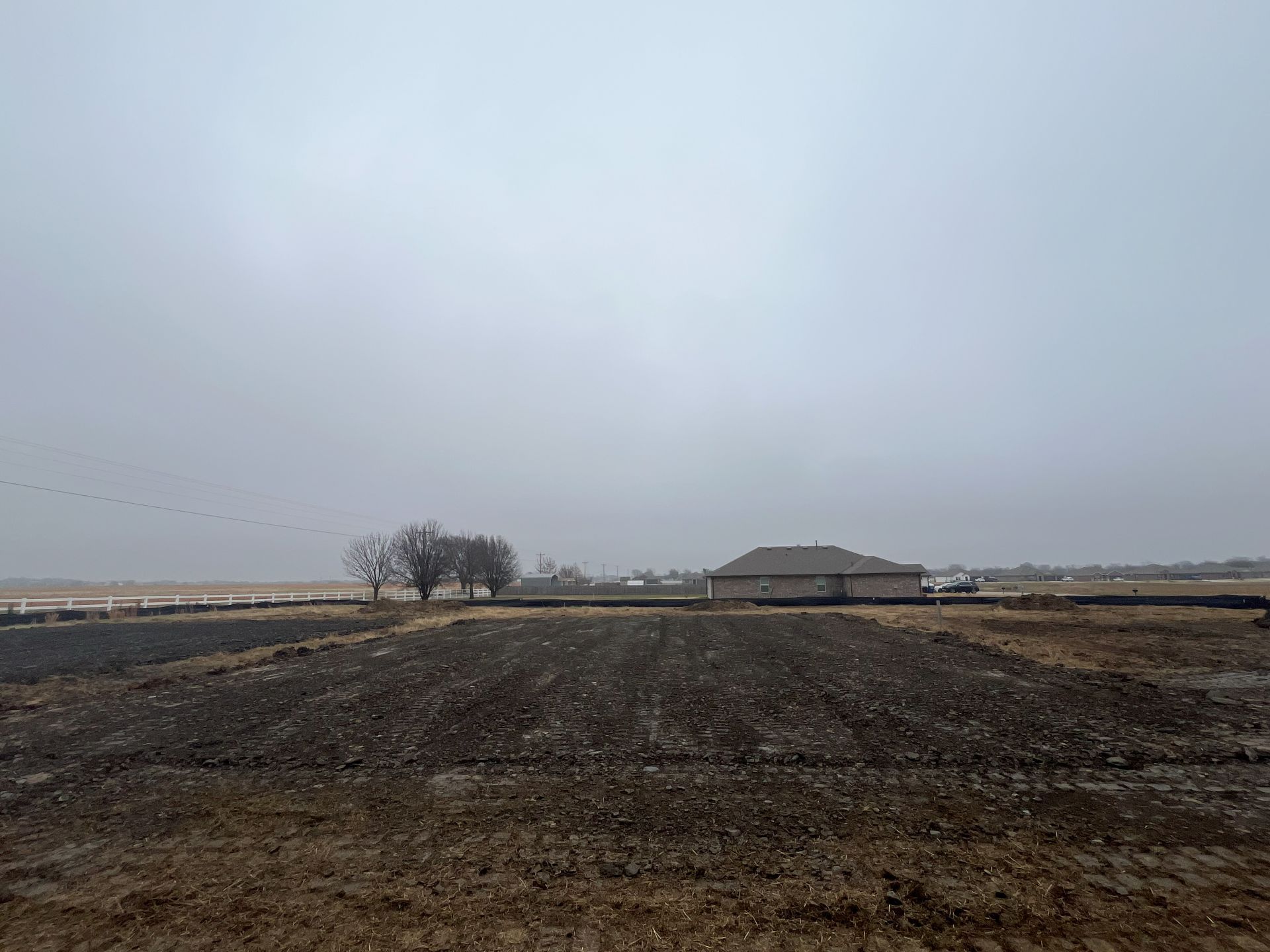 A flat, plowed field under a cloudy, overcast sky, with a house and trees in the distance.
