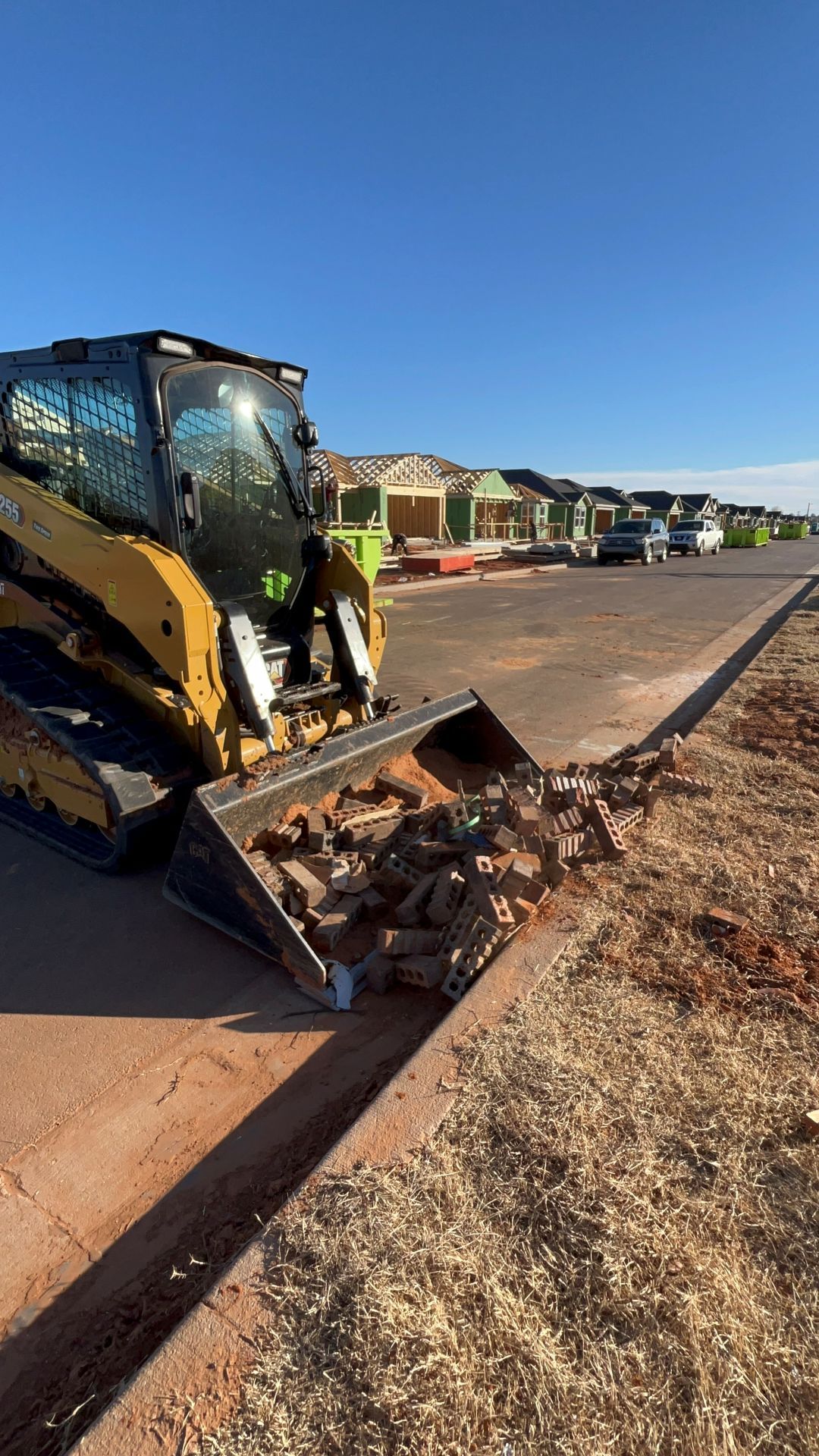 A yellow skid steer loader with a filled bucket sits on a paved street at a construction site with new homes in the back.