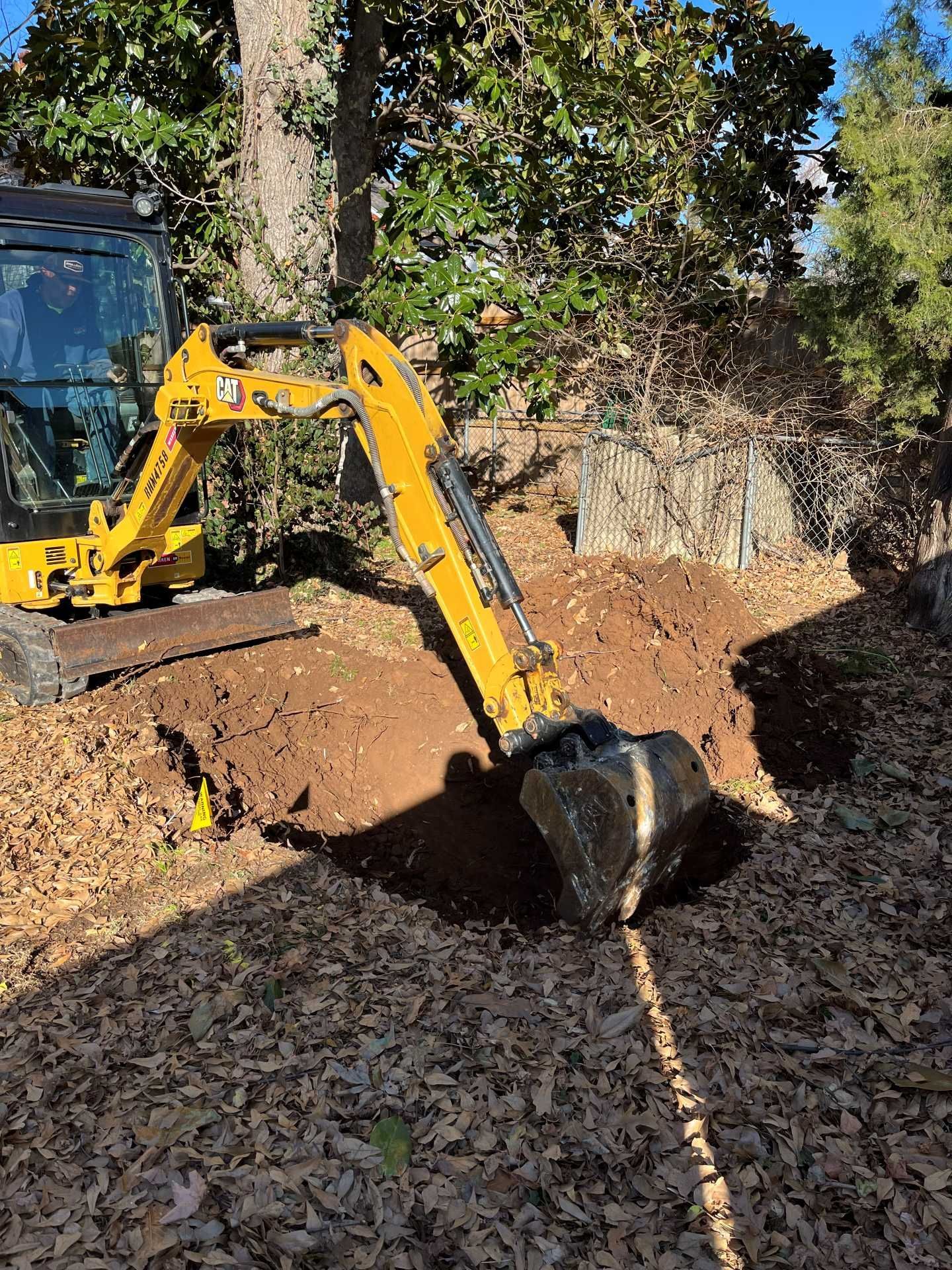 A yellow mini excavator digging a trench in a yard covered with fallen leaves.