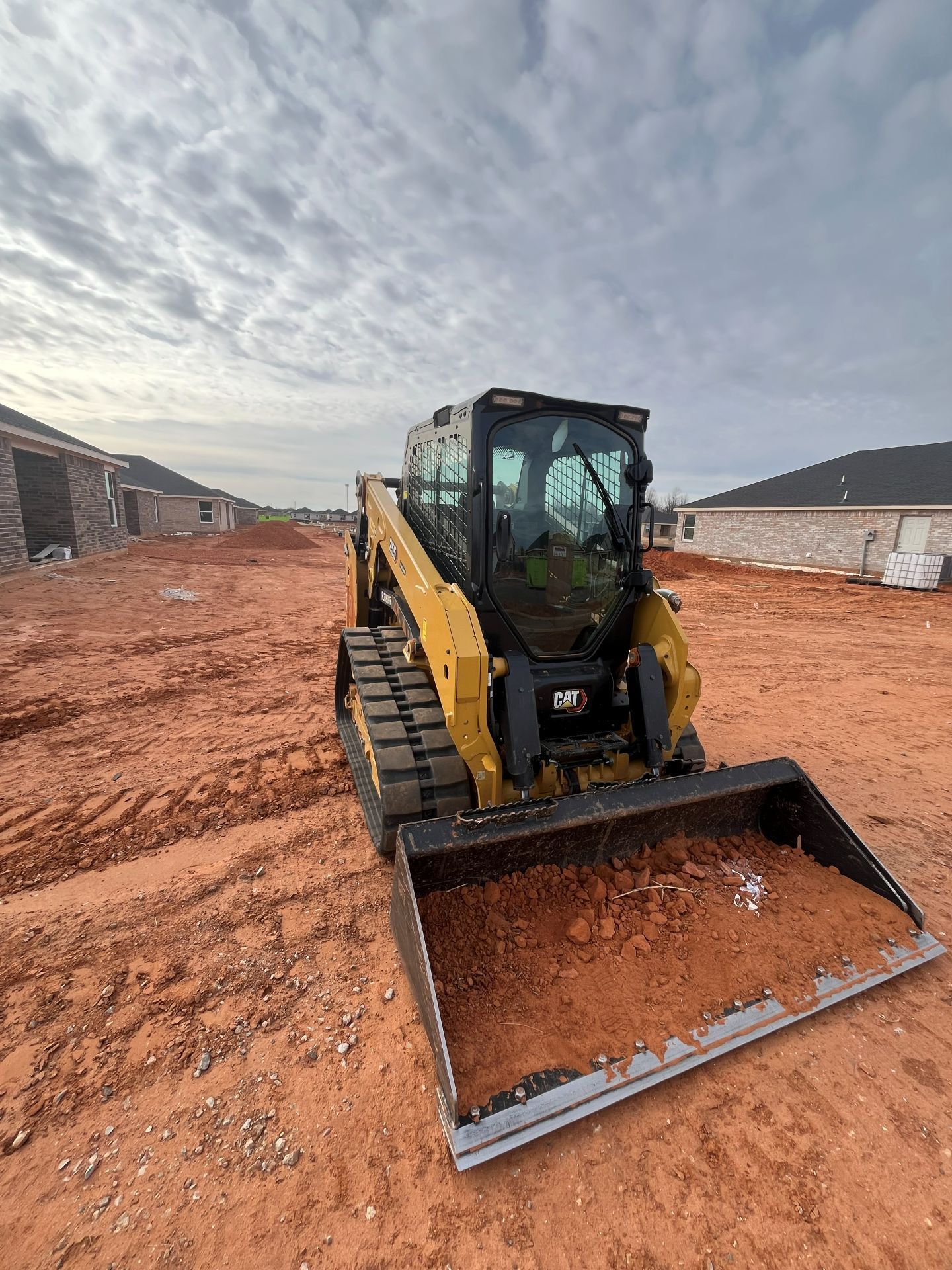 A yellow skid-steer loader with tracks sits on a red dirt construction site between residential houses.