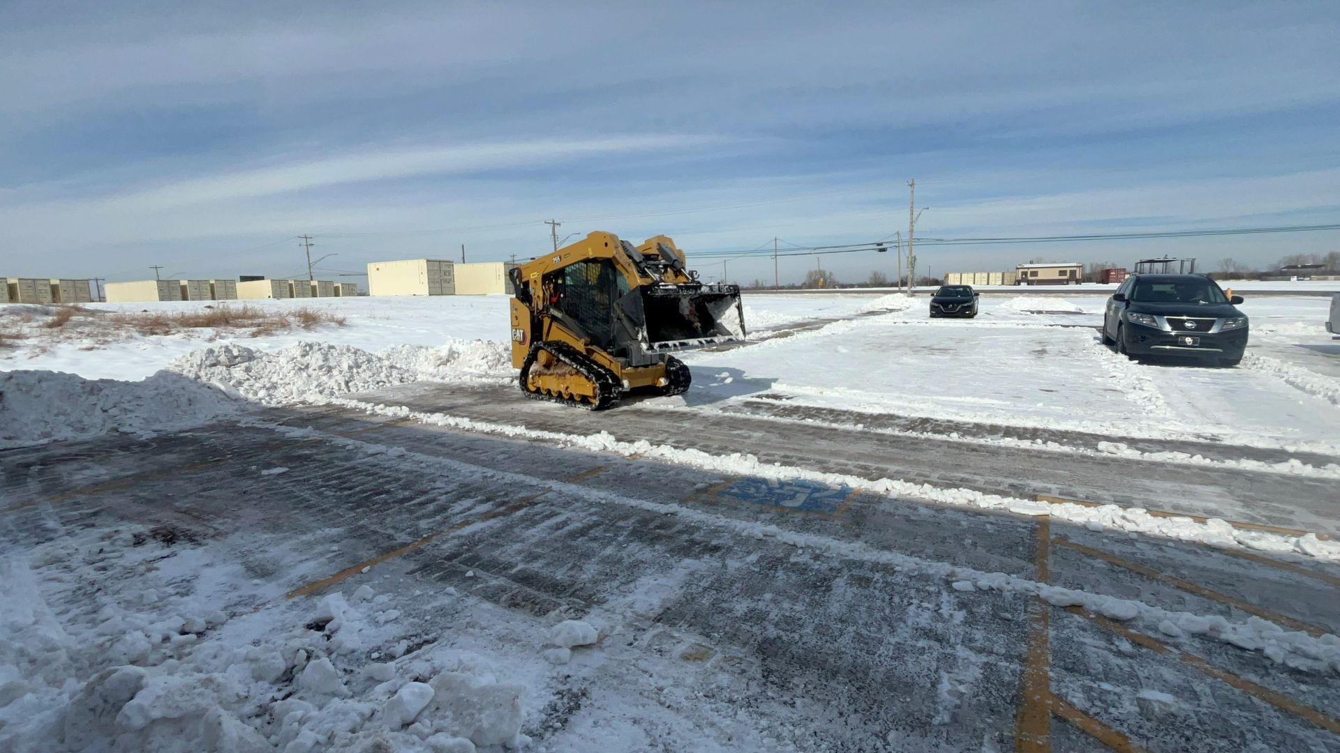 A yellow tracked skid steer clears snow from a parking lot on a sunny winter day.