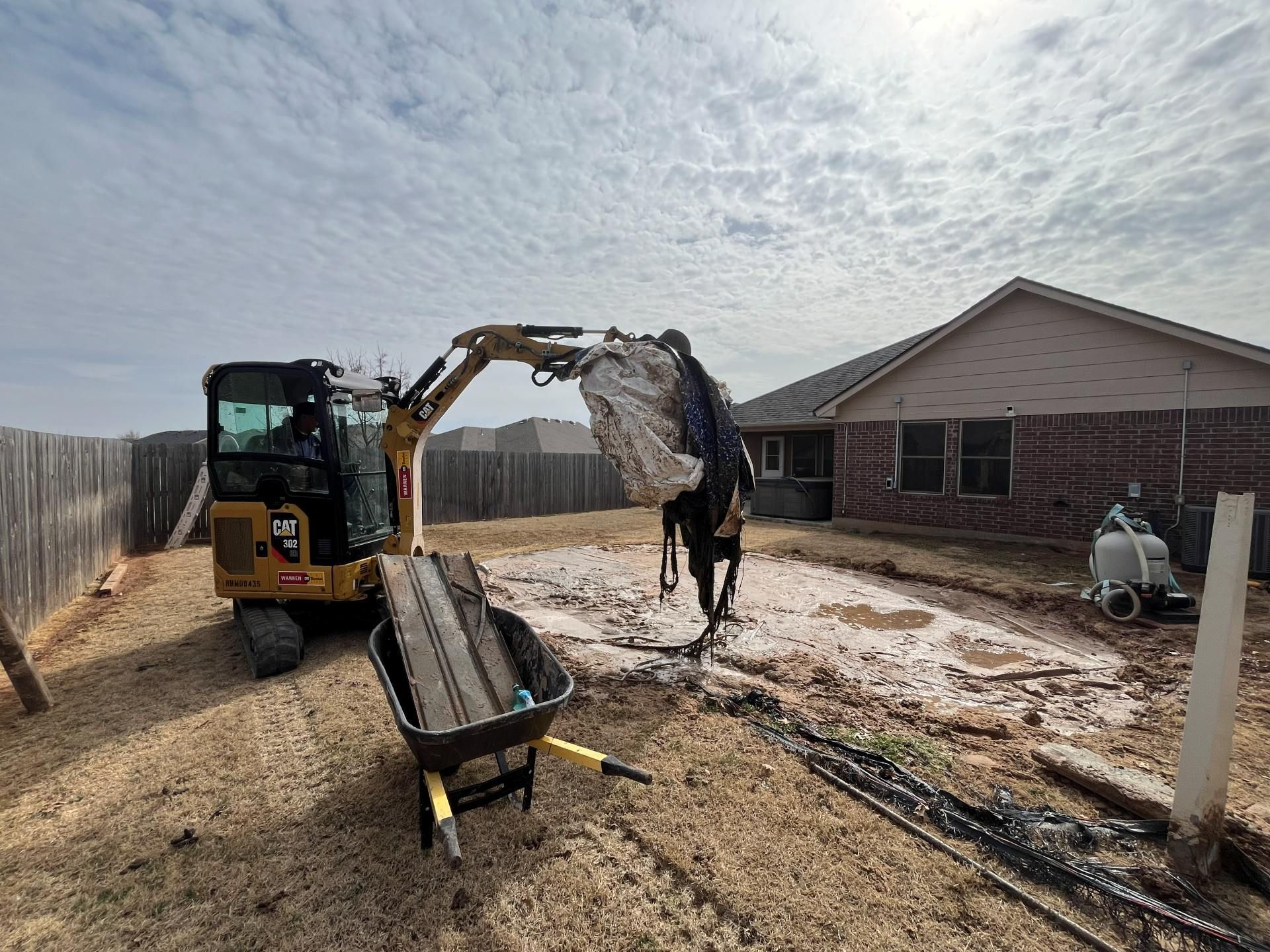 A yellow mini excavator in a backyard lifts a large white construction bag over a dirt-covered area near a brick house.