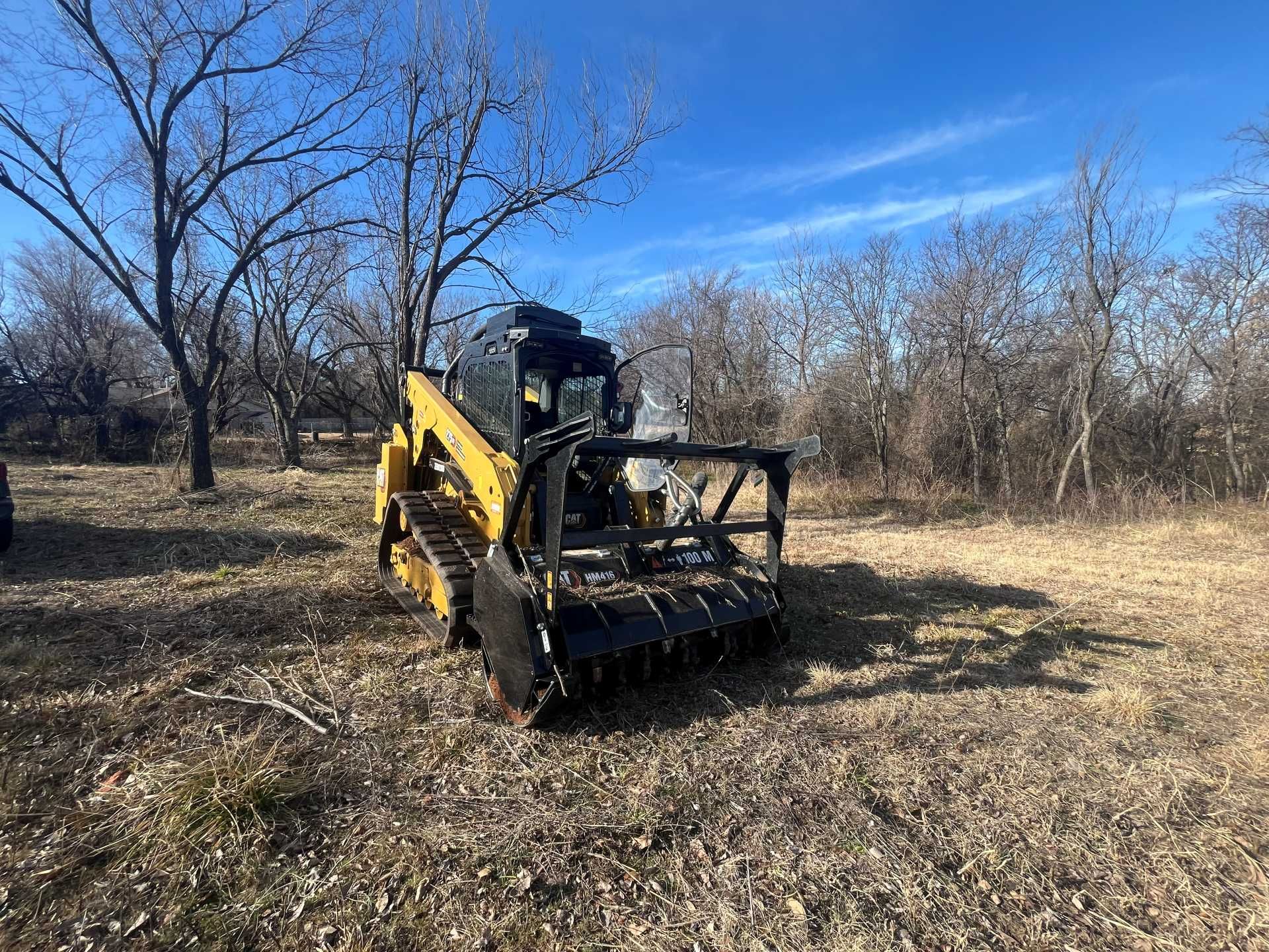 A yellow track skid steer with a forestry mulcher attachment parked in a field with bare trees under a clear blue sky.