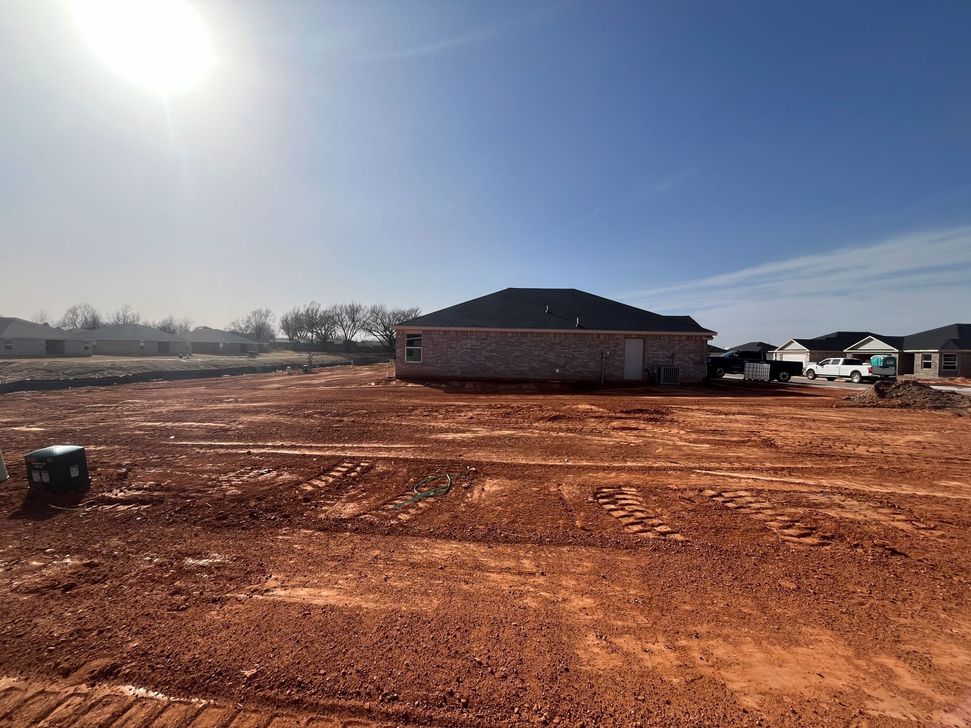 A large, sunny construction site with cleared red earth, featuring a nearly finished brick house in the background.