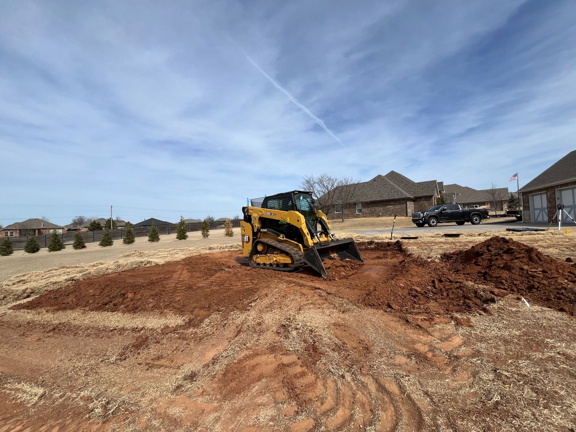 A yellow Caterpillar skid steer loader moves red earth in a residential neighborhood under a bright blue sky.