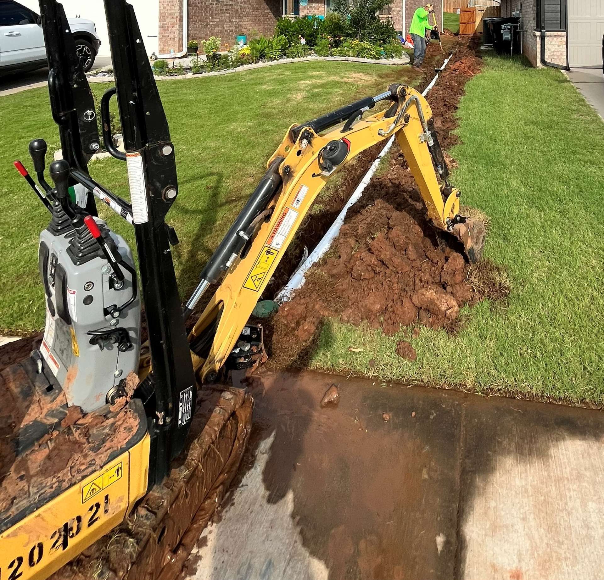 A yellow mini excavator digging a trench through a green residential lawn next to a paved driveway.