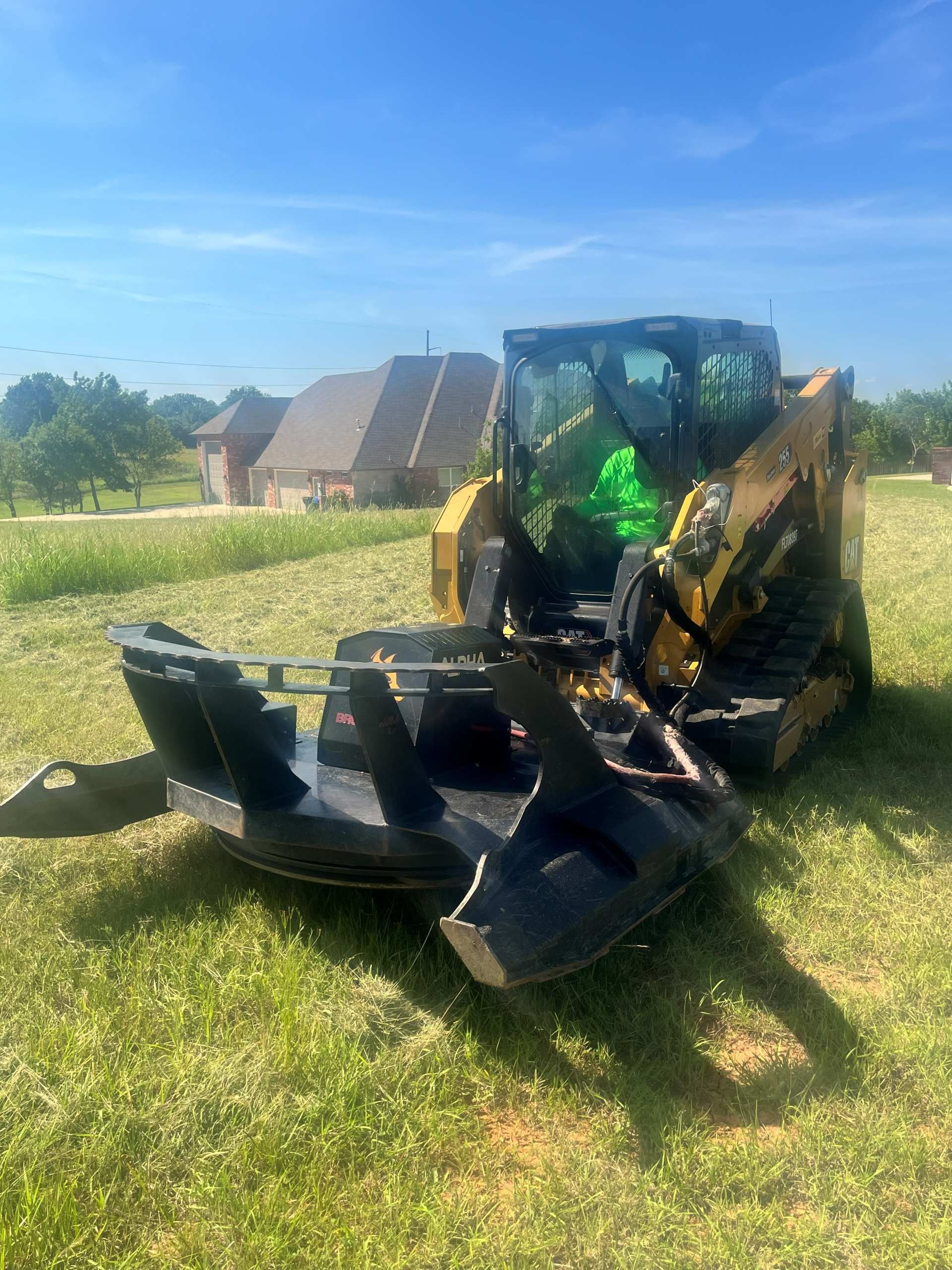 A yellow tracked skid steer with a large black rotary brush cutter attachment parked in a sunny grass field.