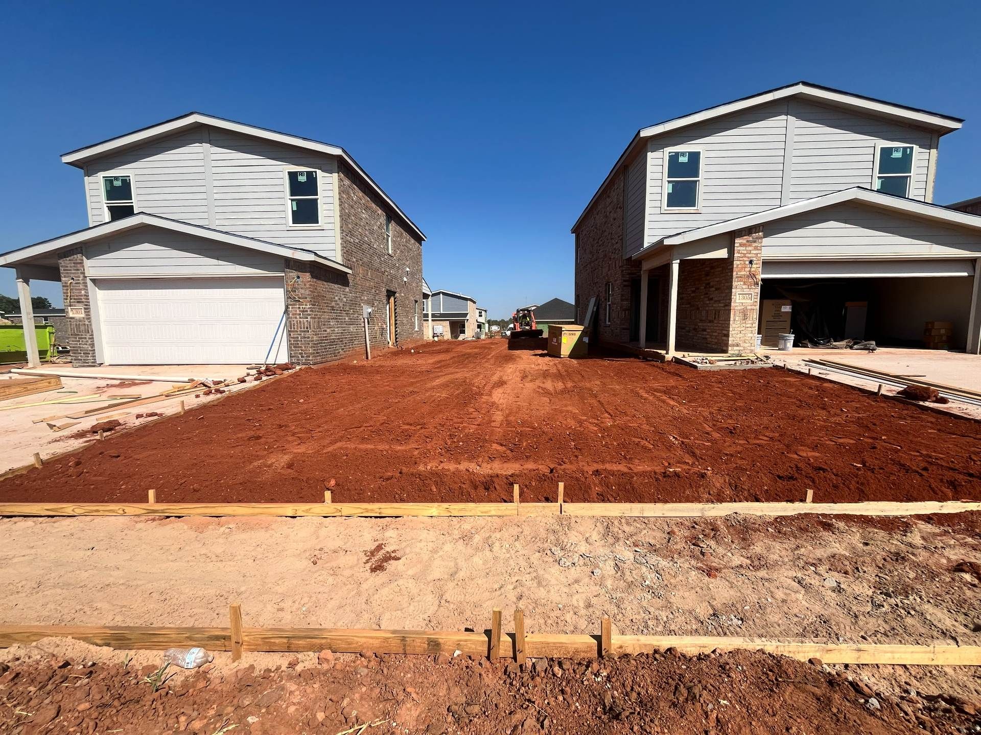 Two newly constructed houses flank a wide patch of prepared red dirt between them, set behind wooden forms for a driveway.