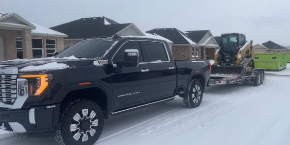 A black pickup truck towing a flatbed trailer with a yellow skid steer loader on a snow-covered suburban street.